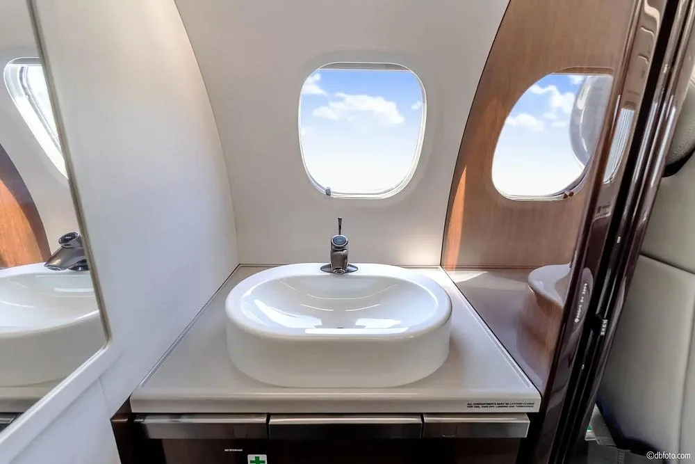 The lavatory area of the private jet, featuring a white sink, faucet, and a window with a view of the sky.