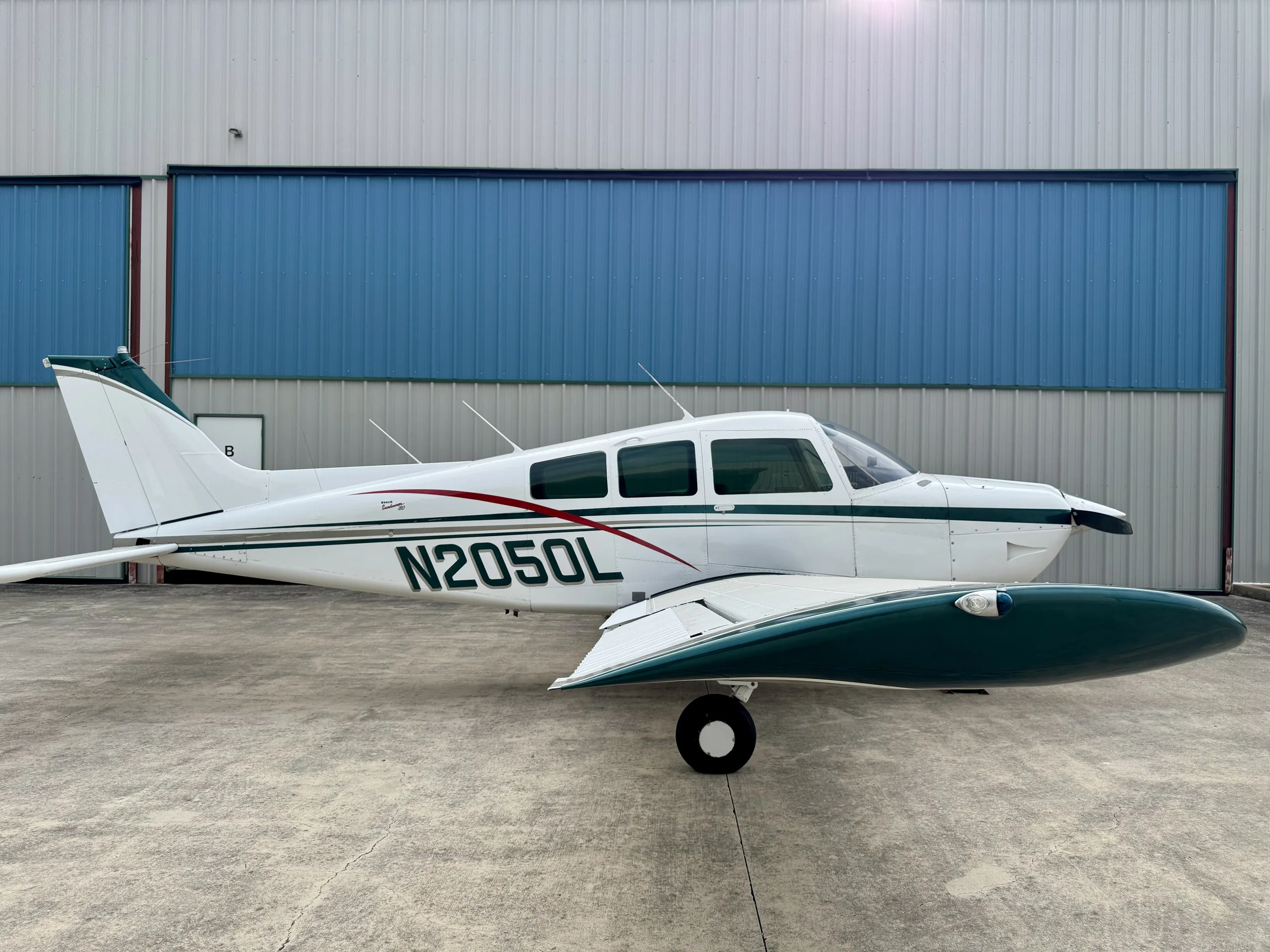 A white and green Beechcraft Sundowner N2050L parked on concrete in front of a hangar.