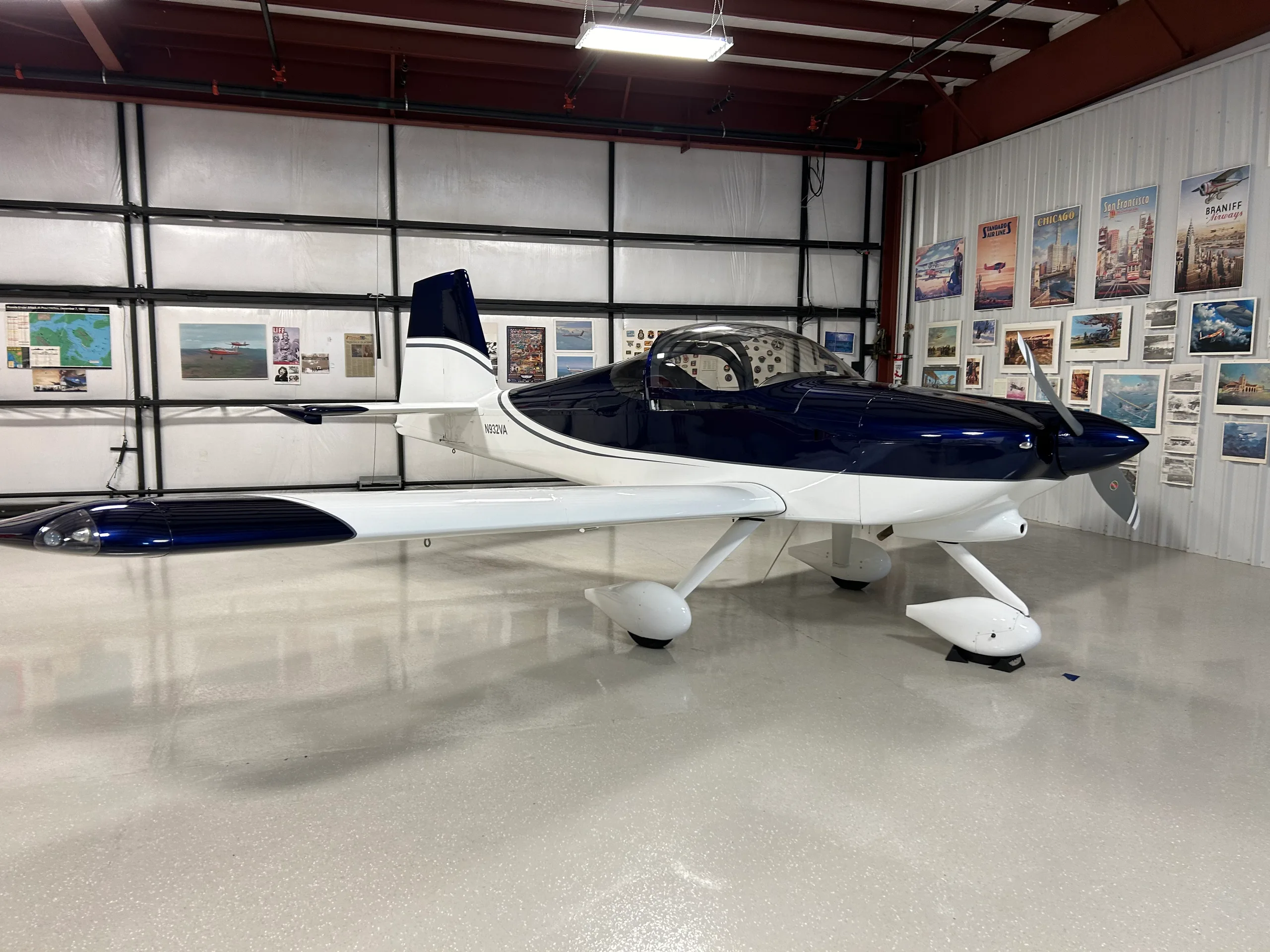 A dark blue and white single-engine aircraft, registration N932VA, parked inside a hangar with aviation posters on the wall.