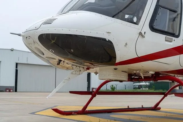 A close-up of the white and red helicopter's nose and red landing skids on a yellow-marked tarmac.