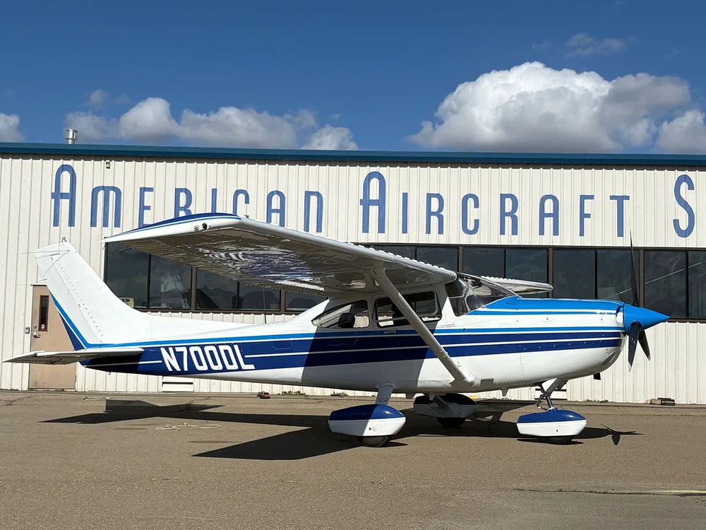 A white and blue Cessna 182 Skylane (N700DL) parked on the tarmac in front of a building.