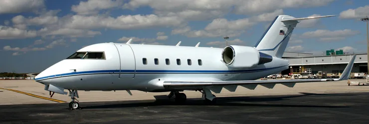 A white Bombardier Challenger business jet with blue stripes parked on an airport tarmac under a partly cloudy sky.