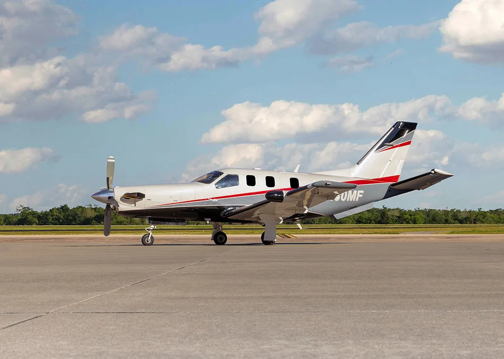 A white and grey TBM 900 series aircraft with red stripes, registration N180MF, parked on an airfield, viewed from the side.
