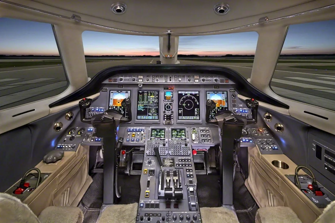 The modern cockpit of a private jet with multiple digital displays and controls, looking out onto a runway at sunset.