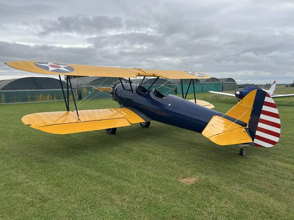 A three-quarter rear view of a blue and yellow biplane with US military insignia on the wings and N60955 registration on the tail, parked on a grassy field.