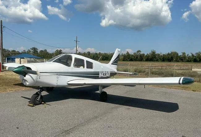 A white Piper Cherokee with green stripes, registration N43348, parked on an asphalt ramp under a partly cloudy sky.