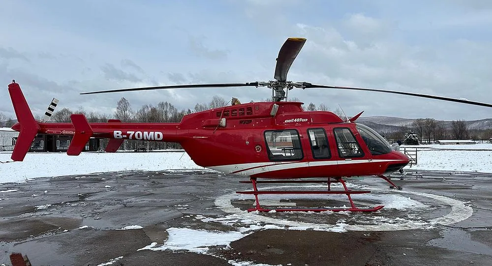 A red Bell 407GXP helicopter with white stripes, tail number B-70MD, parked on a helipad with snow on the ground and distant buildings.