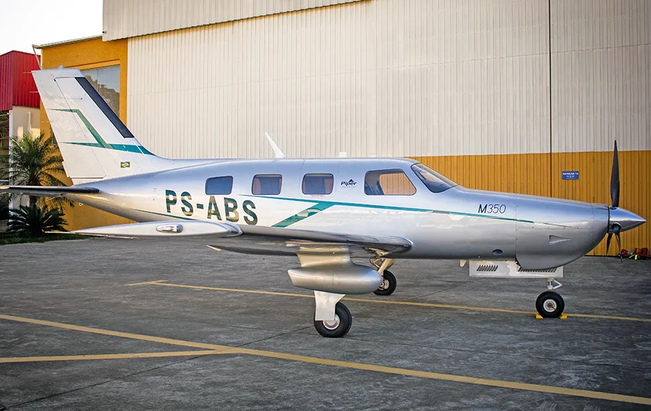 A silver Piper M350 aircraft with green and dark blue stripes is parked on a tarmac in front of a hangar.