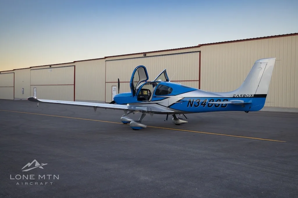 A three-quarter rear view of a blue and silver Cirrus SR22 aircraft with open doors parked on an airfield next to hangars.