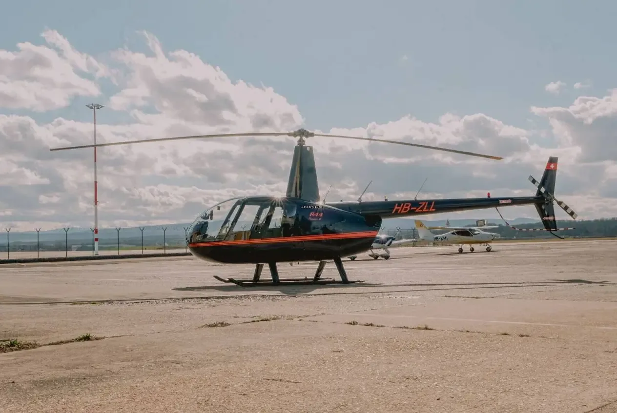 A black Robinson R44 Raven II helicopter, registration HB-ZLL, parked on an airfield with other small aircraft in the distance.