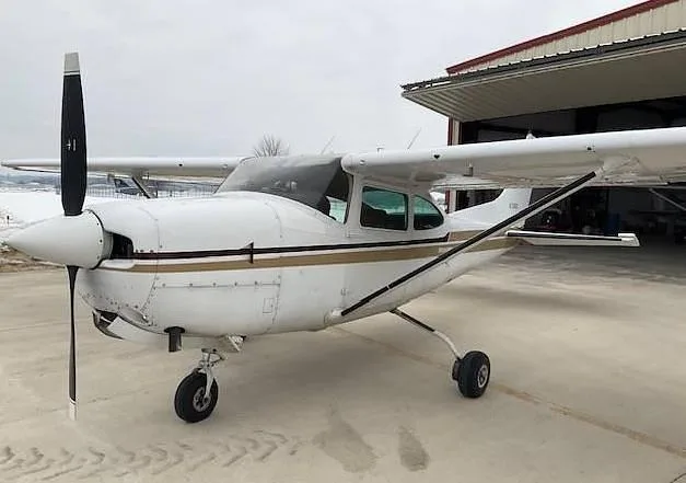 A white Cessna 172 with a gold and black stripe, parked on a tarmac in front of a hangar.