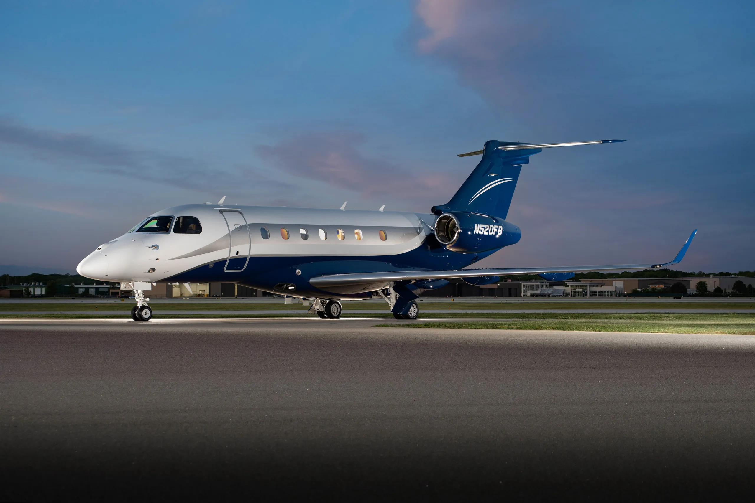 A silver and blue Embraer Phenom 300 business jet, registration N520FB, is parked on a tarmac at dusk with landing lights on.