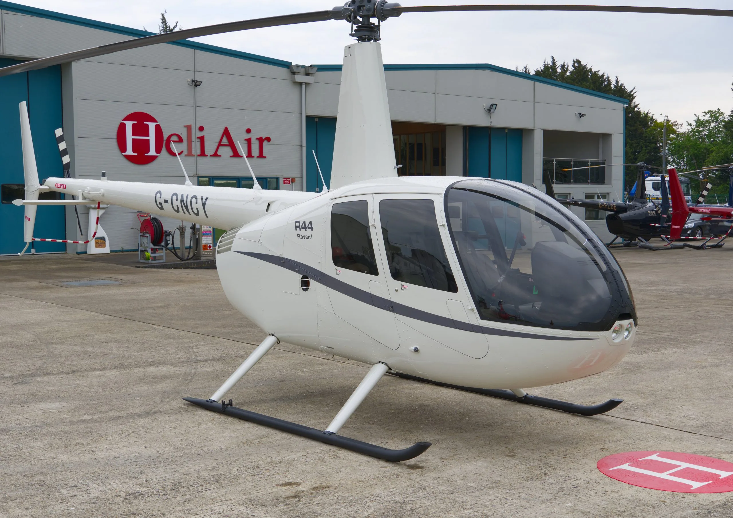 A white Robinson R44 Raven I helicopter (G-GNGY) is parked on a tarmac in front of a HeliAir hangar.