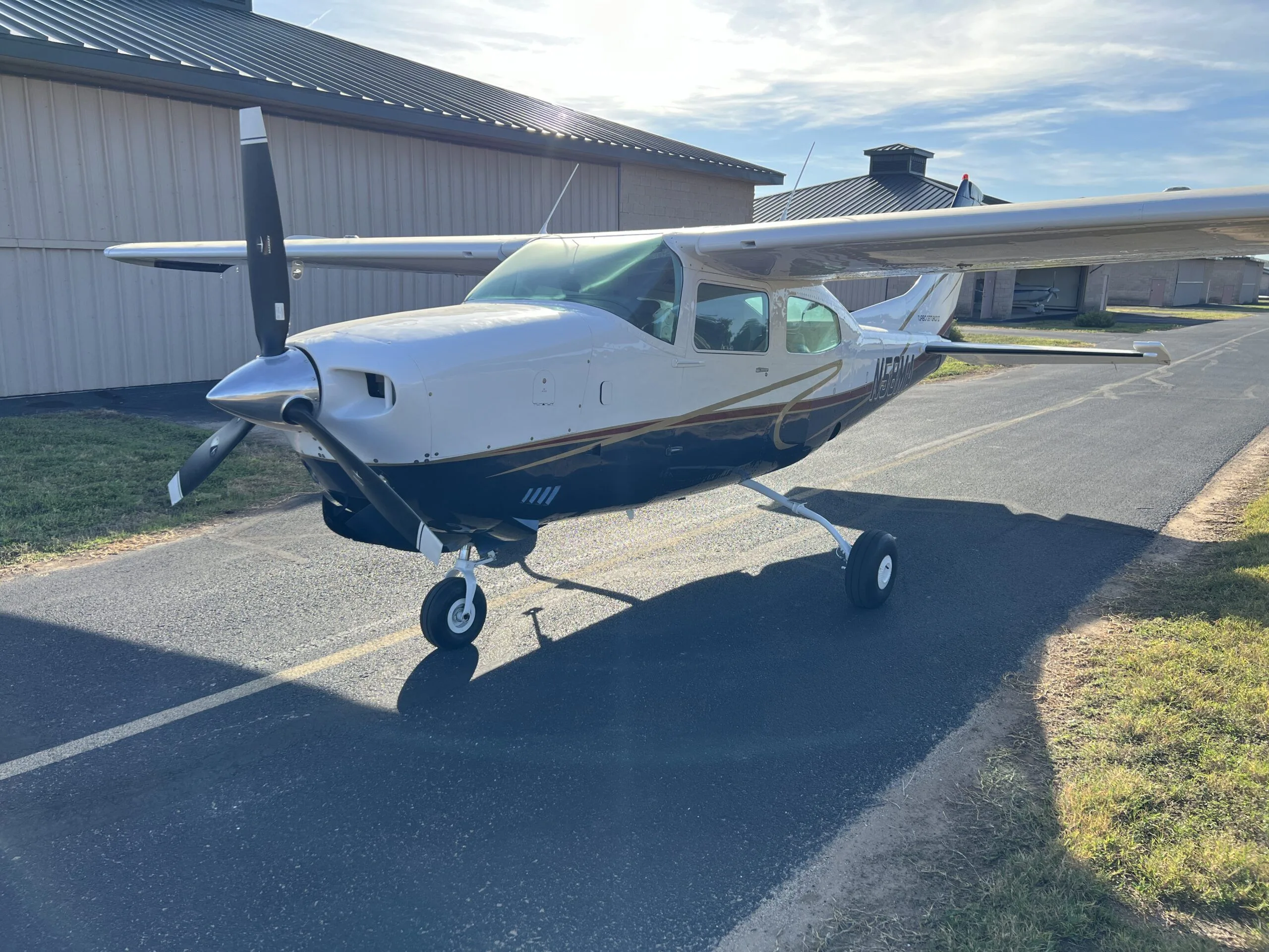 A white and blue Cessna 210, N58MA, with gold trim, parked on an asphalt taxiway next to a hangar.