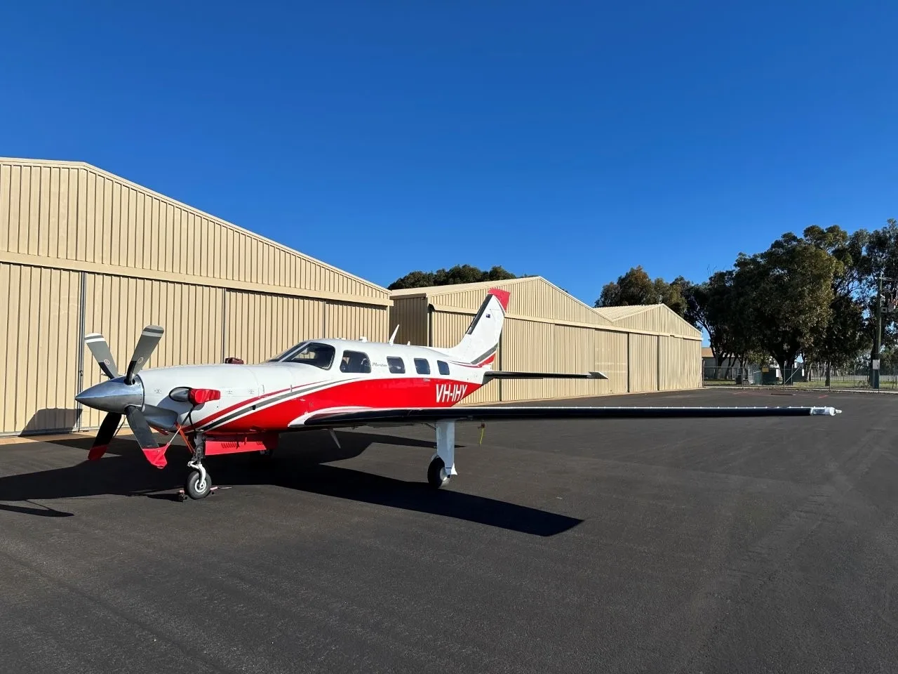 A white and red Piper Meridian aircraft with registration VH-IHY parked on a tarmac next to hangars under a clear blue sky.