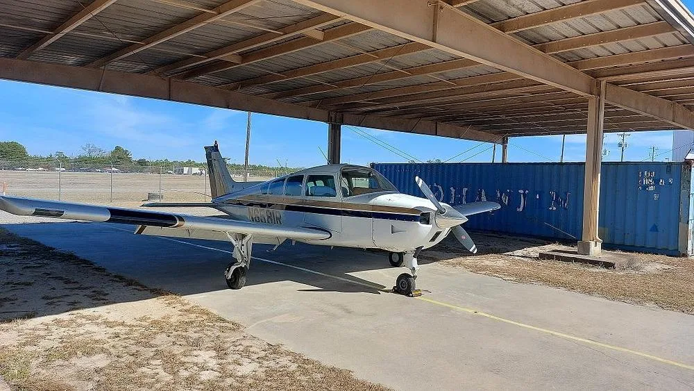 A white and gold single-engine propeller aircraft (Cessna 177 Cardinal) with registration N6581R is parked under an open-sided hangar, viewed from the front-right.