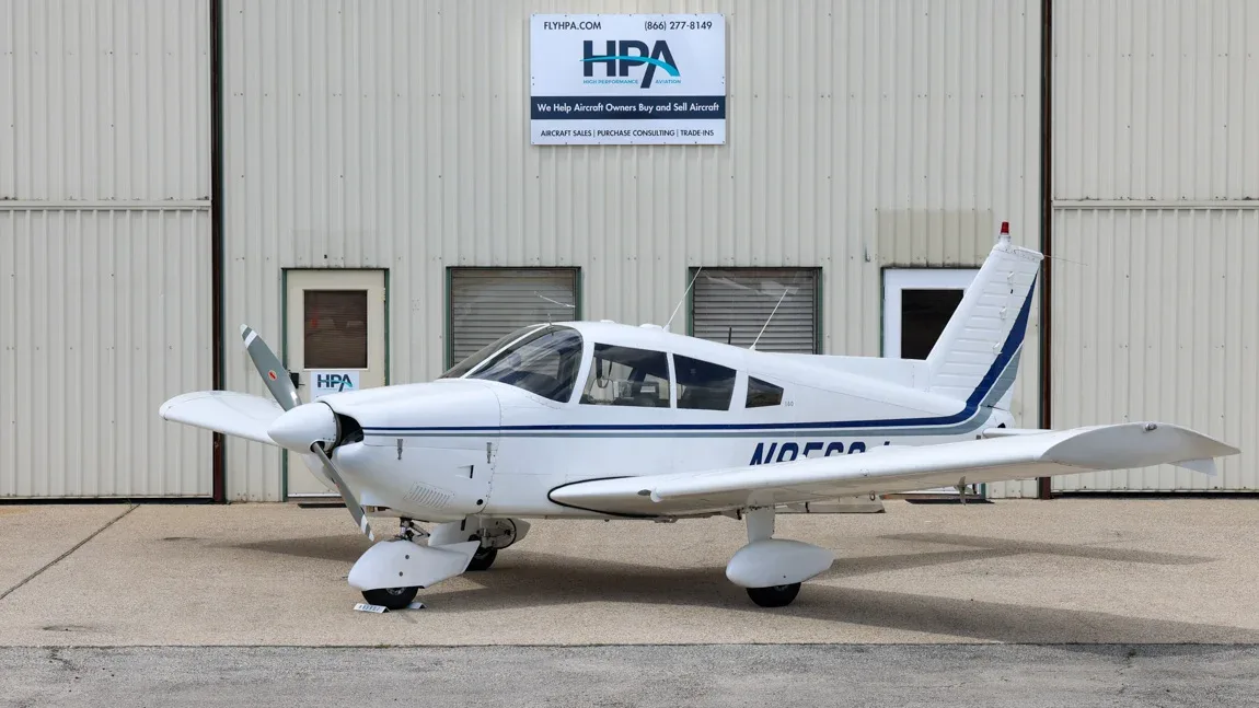 A white single-engine propeller aircraft with blue and grey stripes is parked on an tarmac in front of a hangar.