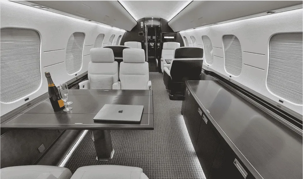A private jet cabin interior with light-colored seats, a table with a laptop and champagne, and dark wood accents.