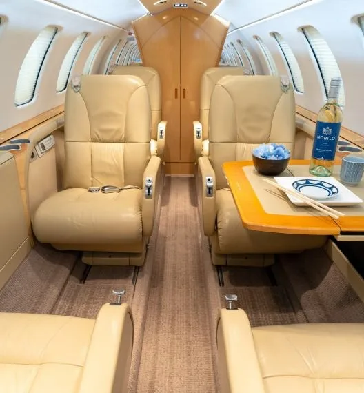 The interior cabin of a business jet features tan leather seats, a table set with a bottle of wine, and a bowl.