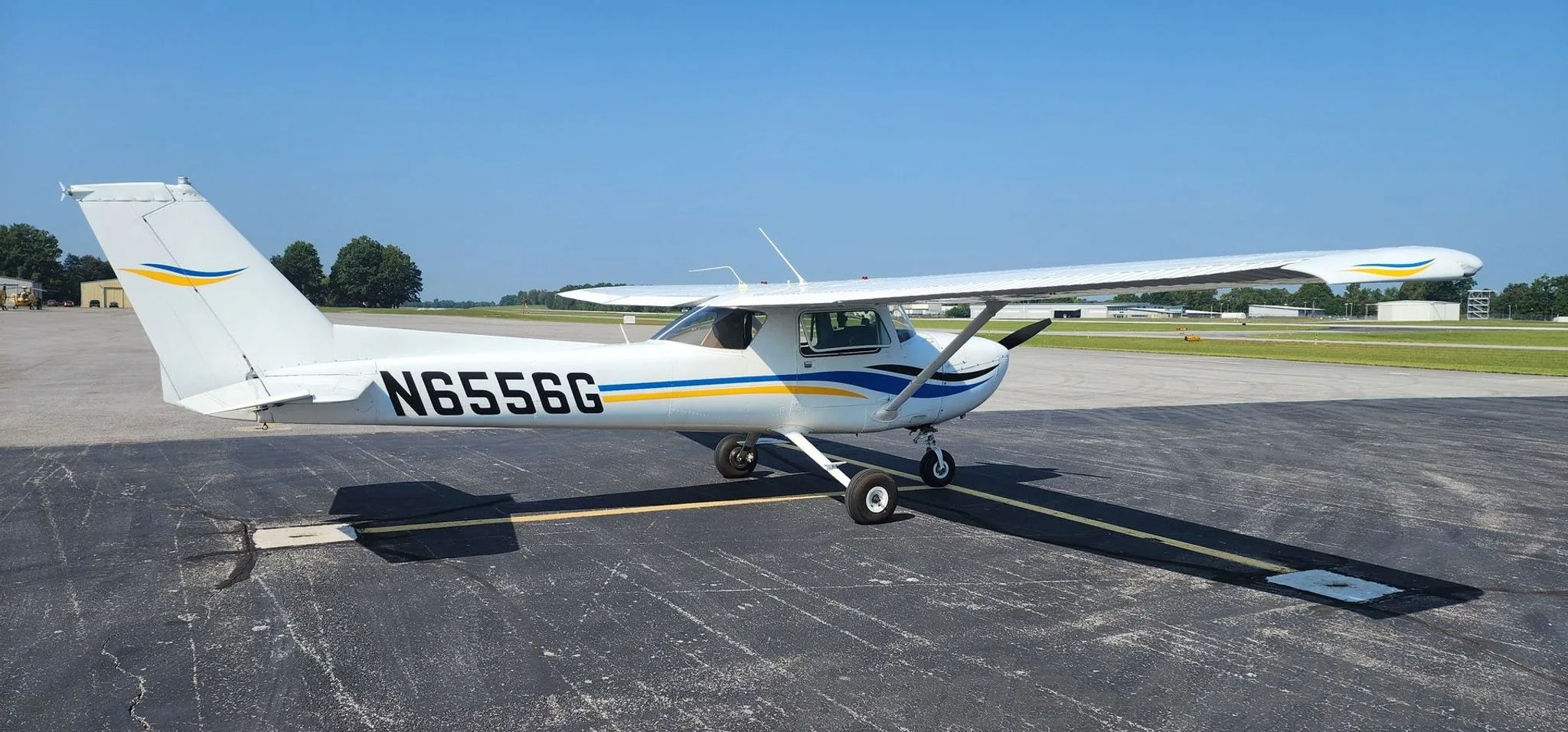 A white Cessna 150 with blue and yellow stripes on the tail and fuselage, parked on an asphalt tarmac under a clear blue sky.