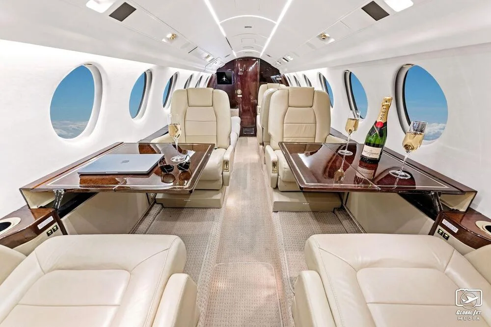 The luxurious interior of a business jet cabin with cream leather seats, two tables set with champagne, and large oval windows showing clouds.