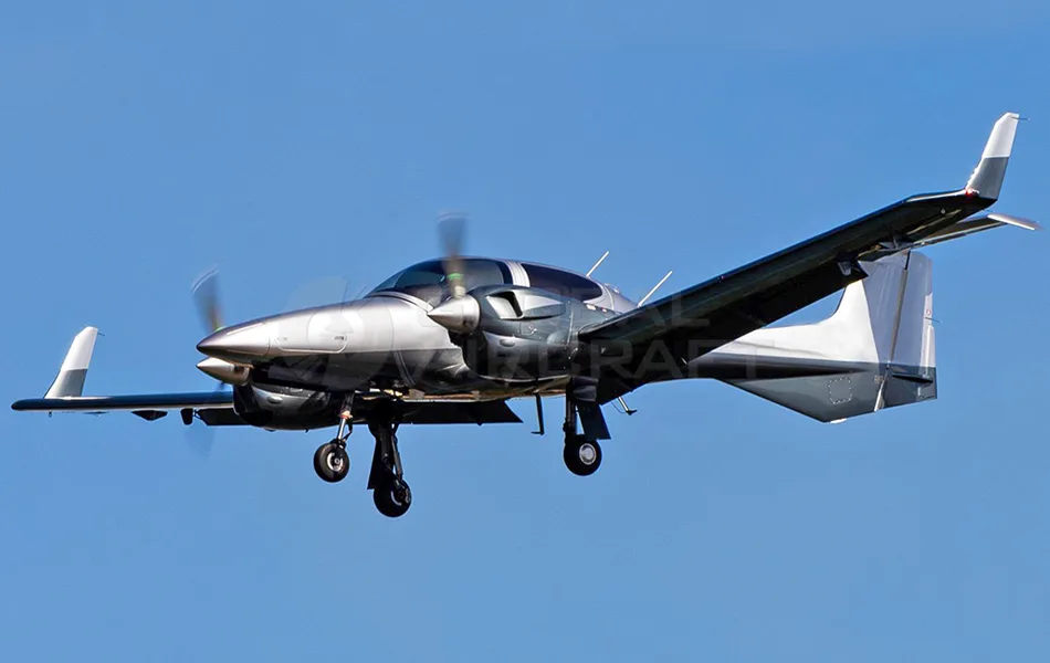 A silver and grey twin-engine Diamond DA62 aircraft with landing gear down, flying against a clear blue sky.