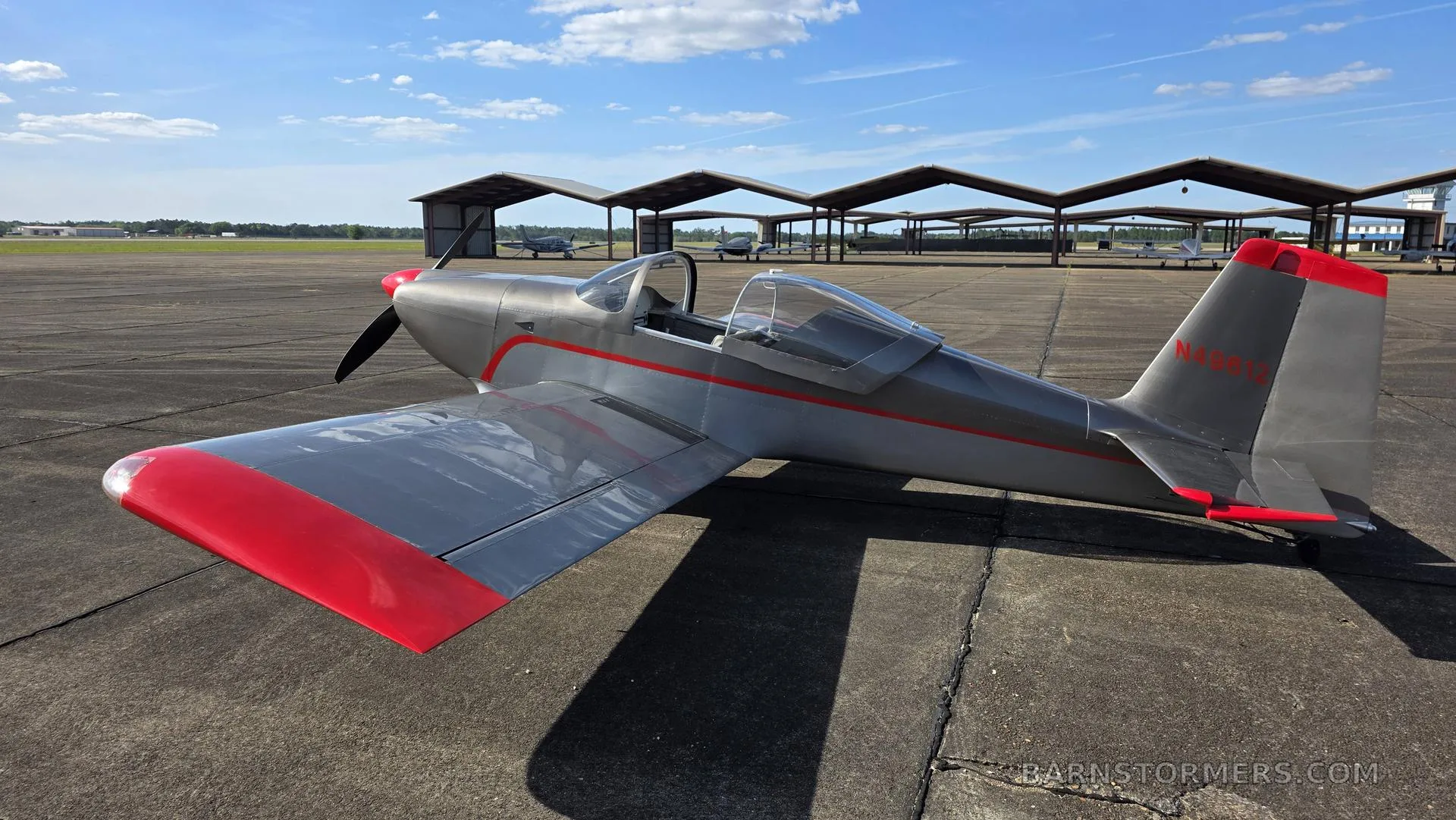 A silver single-engine aircraft with red wingtips and tail accents, parked on an asphalt tarmac with open hangars in the background under a blue sky.