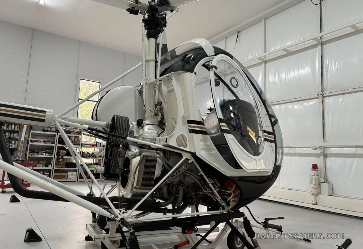 A close-up view of the engine and fuselage of a black and white Schweizer 300CBi helicopter inside a hangar.
