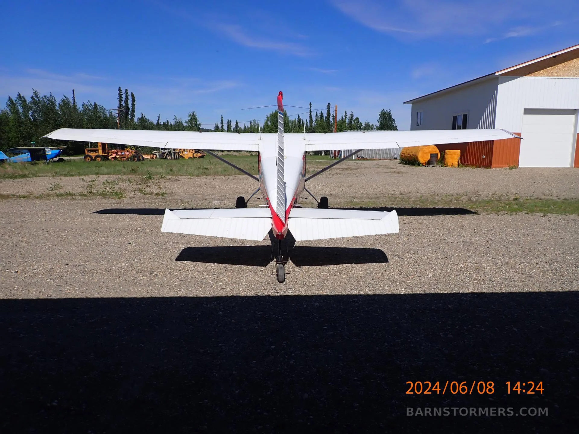 A rear view of a red and white Cessna 180 aircraft parked on a gravel lot with hangars in the background.