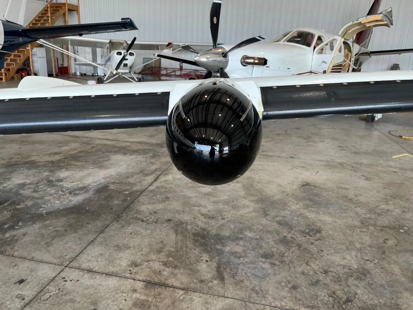 A close-up view of a black pod on the wing of a white and blue aircraft inside a hangar.