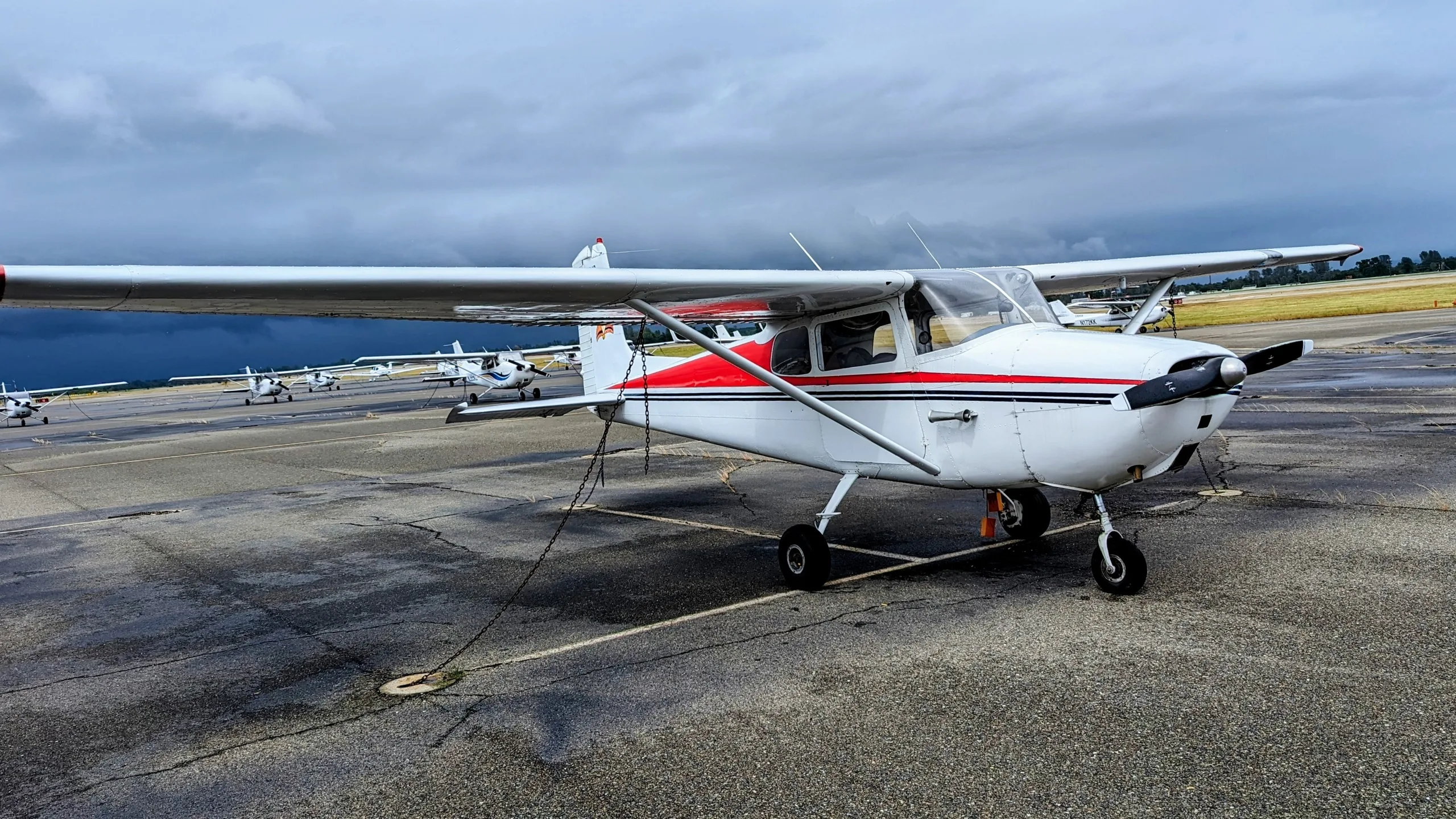 A white 1960 Cessna 172A with red and black stripes is parked on a wet tarmac under a cloudy sky.