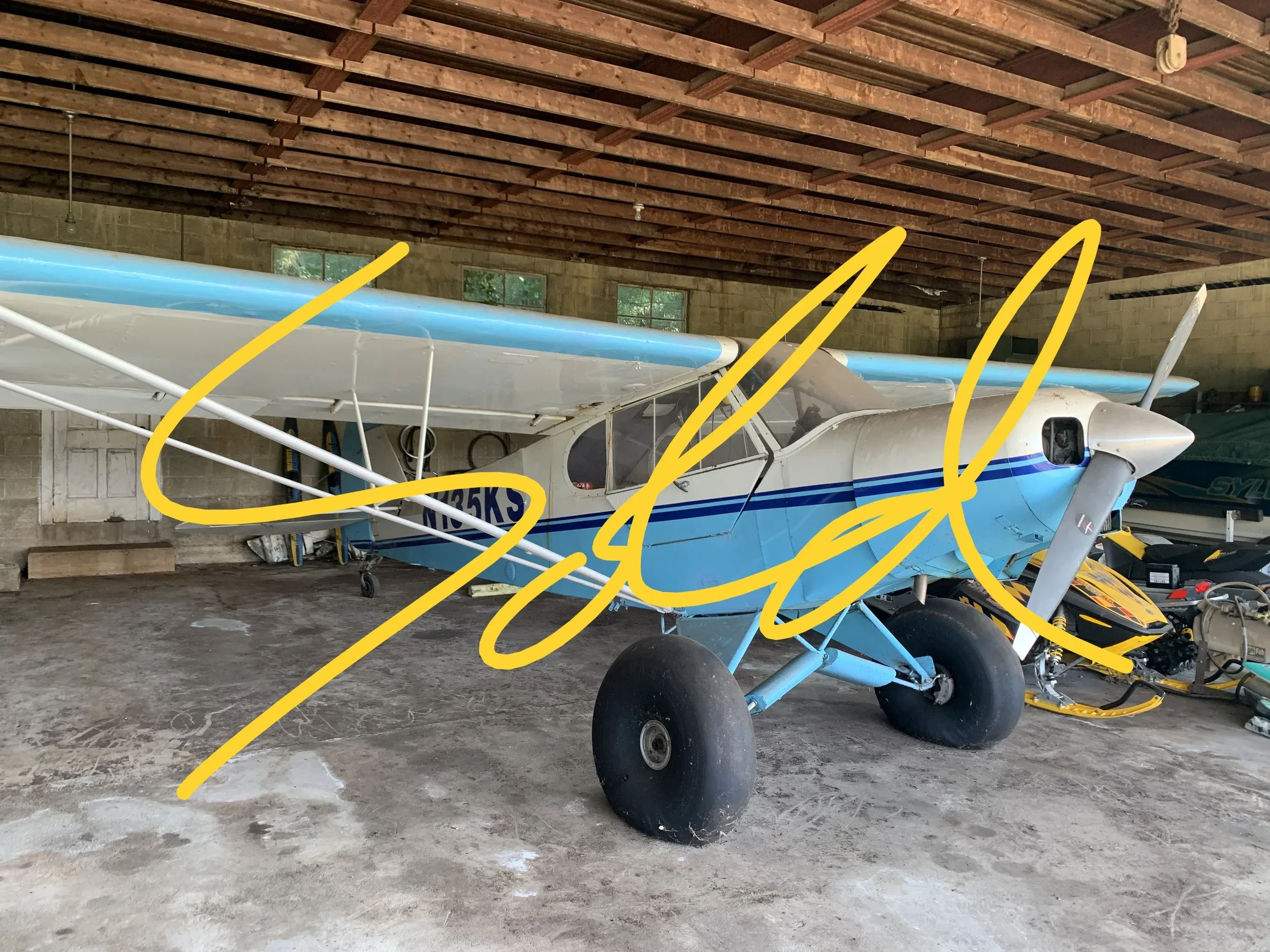 A light blue and white high-wing monoplane with large tundra tires is parked inside a rustic hangar.