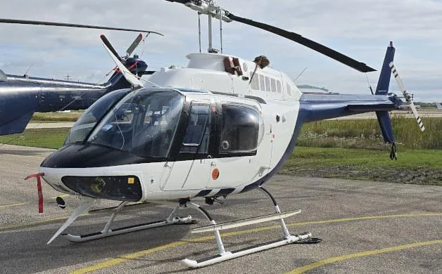 A white and blue Bell 206 helicopter is parked on an asphalt tarmac with another aircraft in the background.