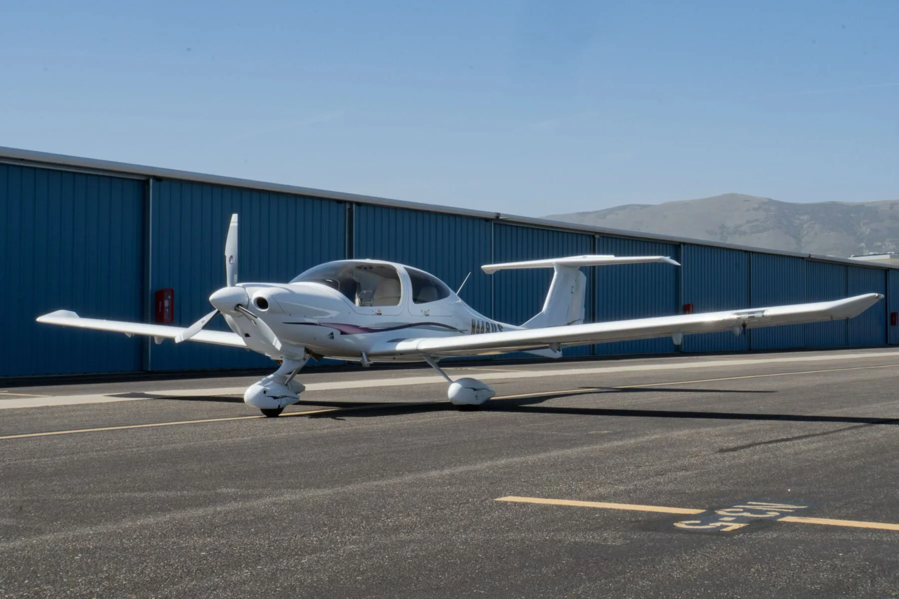 A white Diamond DA40 Star XL aircraft with purple and grey stripes, registration N448US, parked on an tarmac in front of a blue hangar.