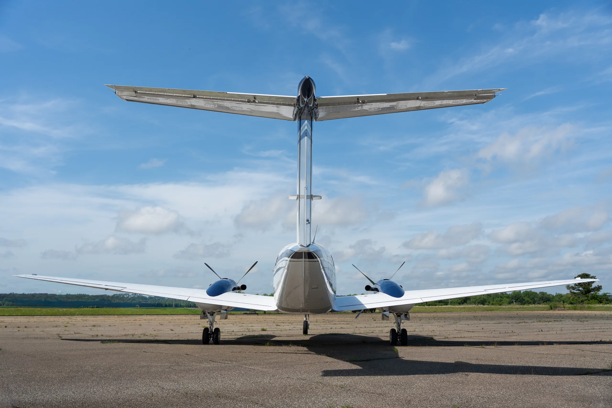 A rear view of a white and blue Beechcraft King Air twin-turboprop aircraft with a T-tail on a tarmac.
