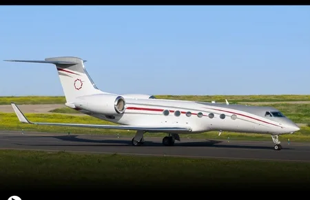 A white Gulfstream G550 business jet with red and grey stripes on a runway with green fields in the background.