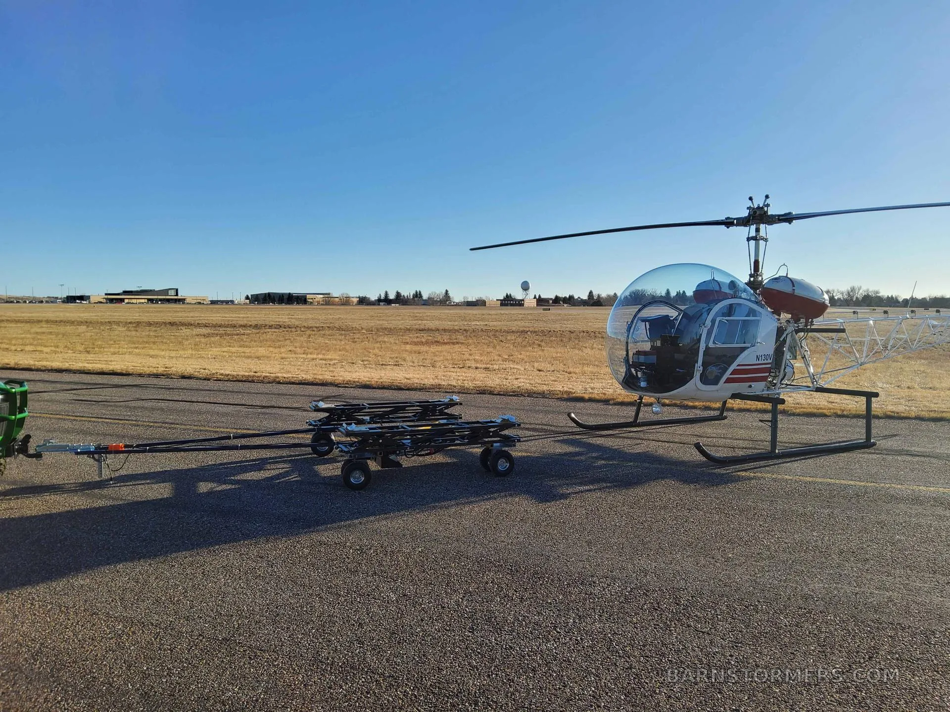 A white helicopter with a clear bubble cockpit and red accents, parked on an airfield next to a ground handling cart.