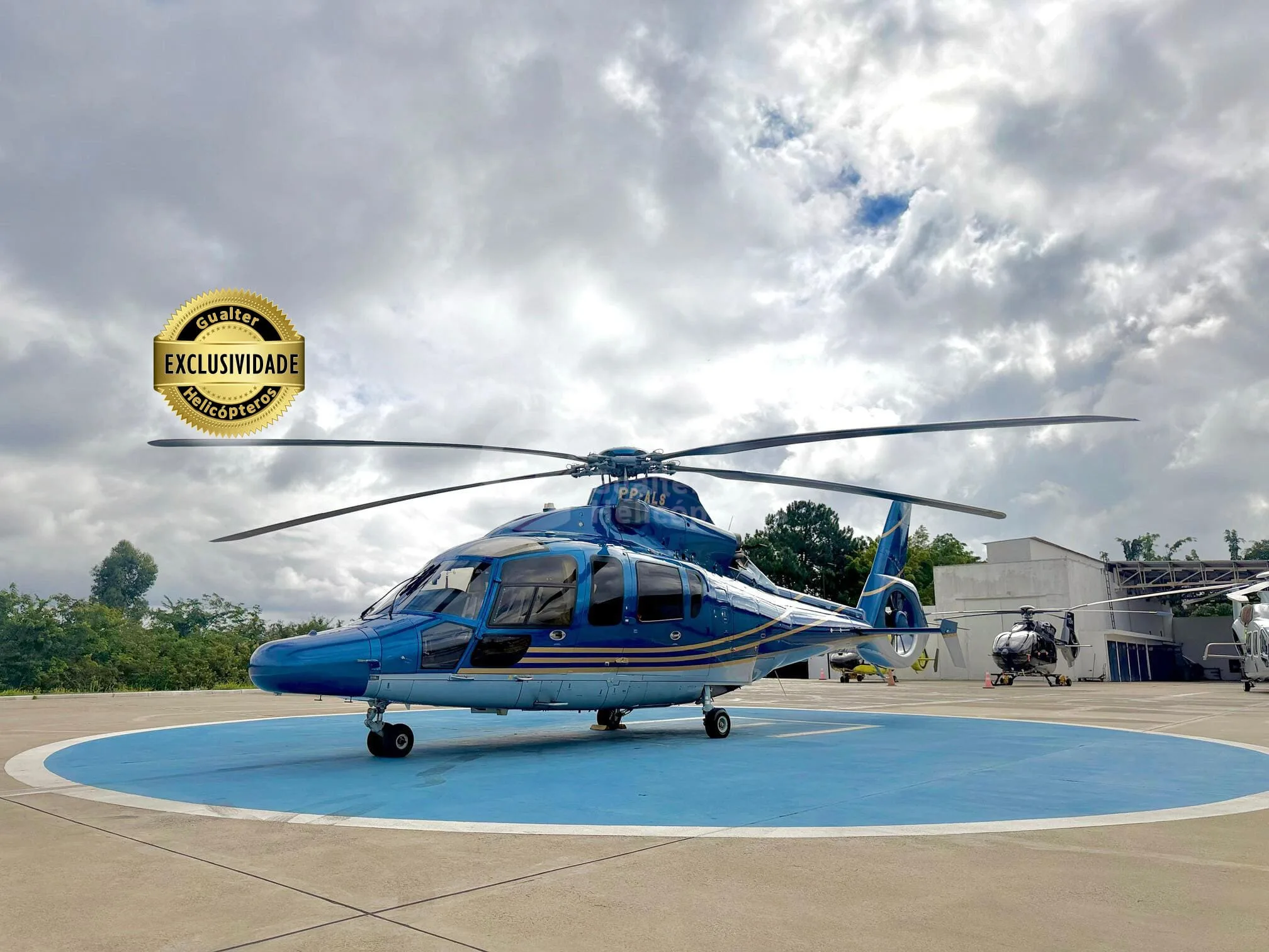 A blue and silver AgustaWestland AW109 helicopter parked on a blue circular helipad under a cloudy sky.