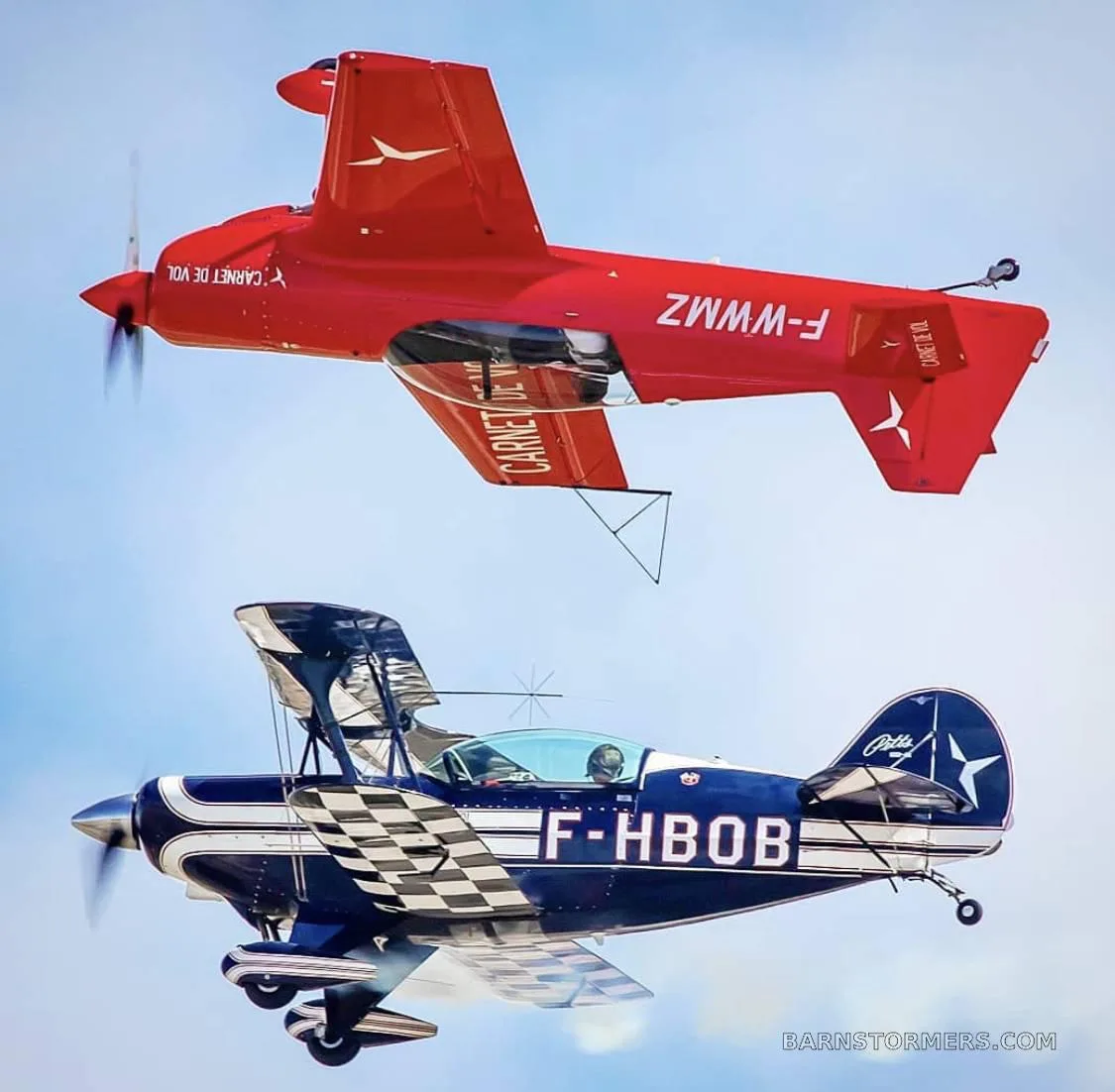 A red single-wing aerobatic aircraft (F-WWMZ) flies above a blue biplane (F-HBOB) with a checkered pattern, both in the sky.