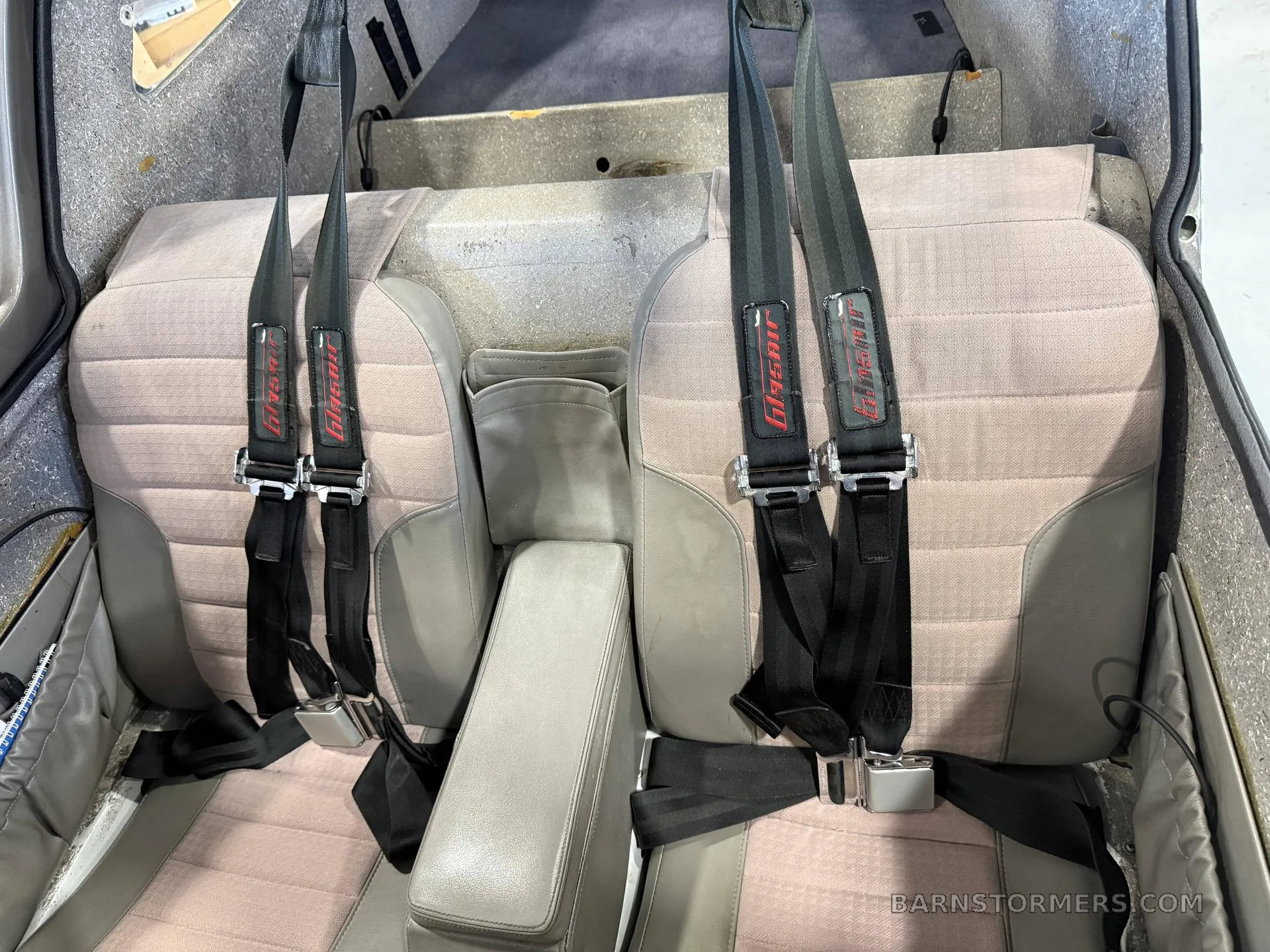 The aircraft's interior shows two light pink fabric rear seats with black four-point harnesses and a gray center console.