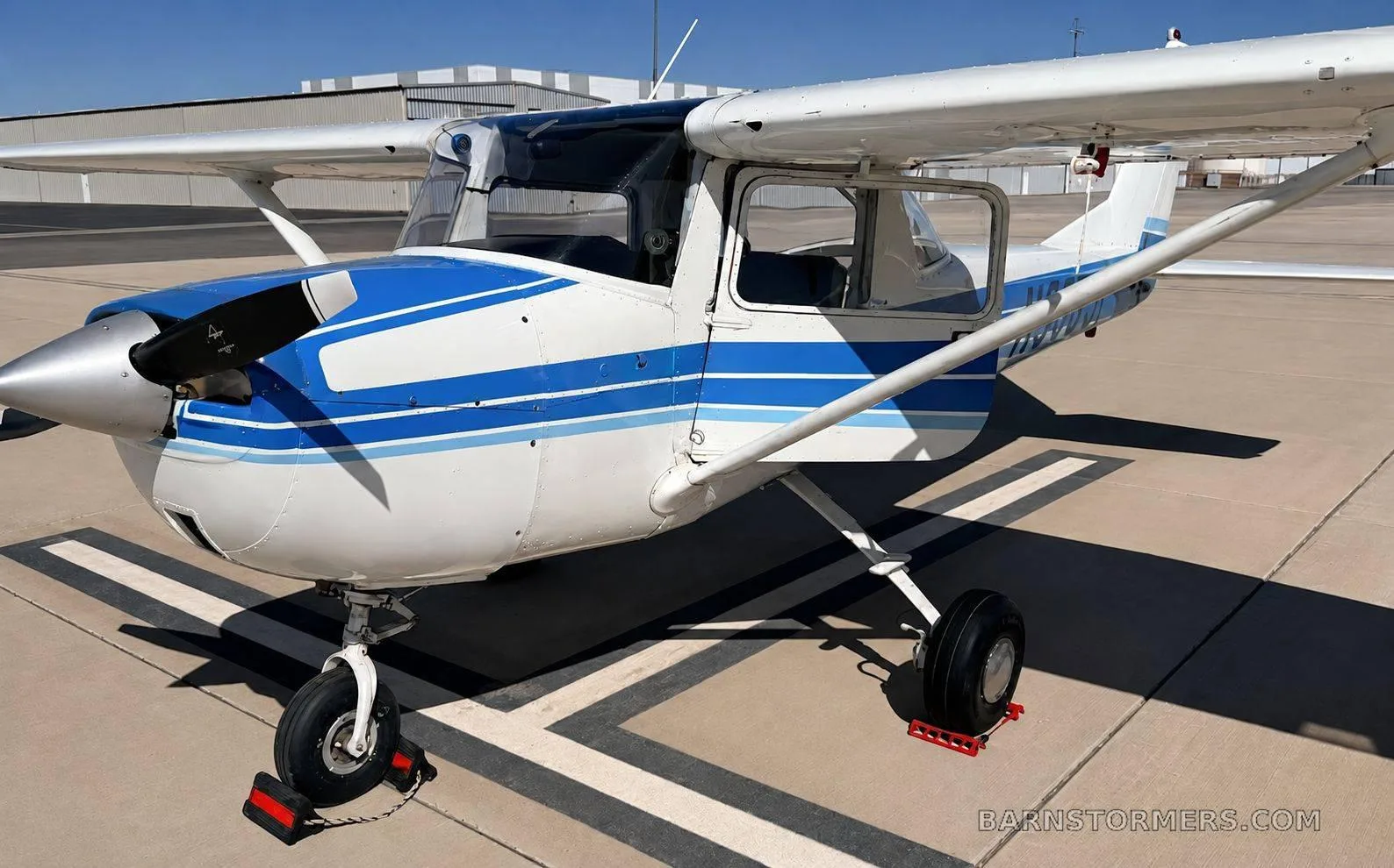 A side-front view of a white and blue single-engine propeller aircraft, with wheel chocks, parked on an airfield.