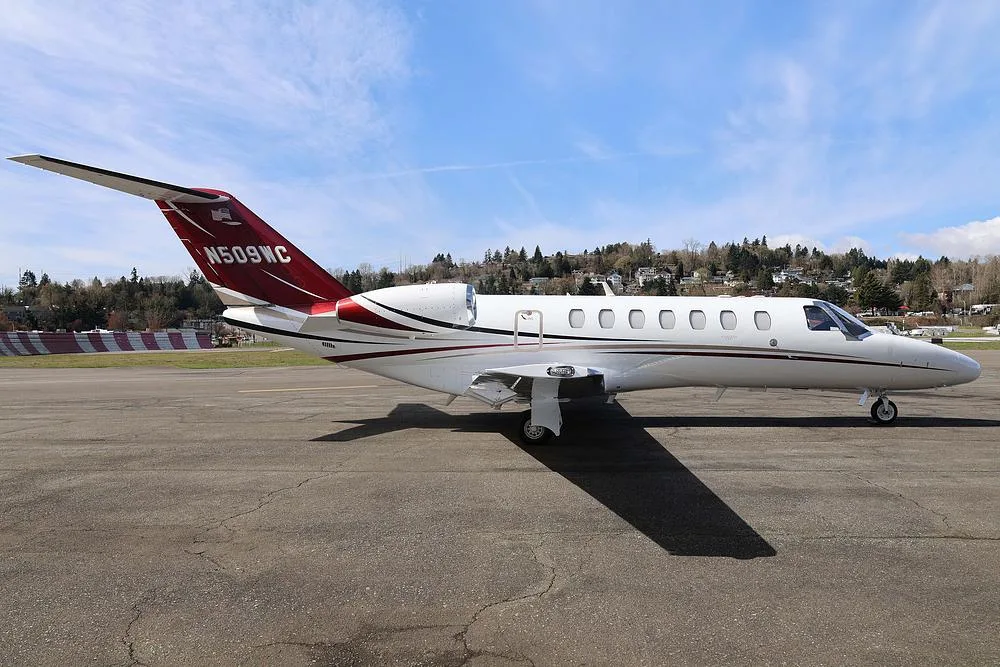 A white private jet with red and black accents, tail number N509WC, parked on an tarmac with a wooded hillside in the background.