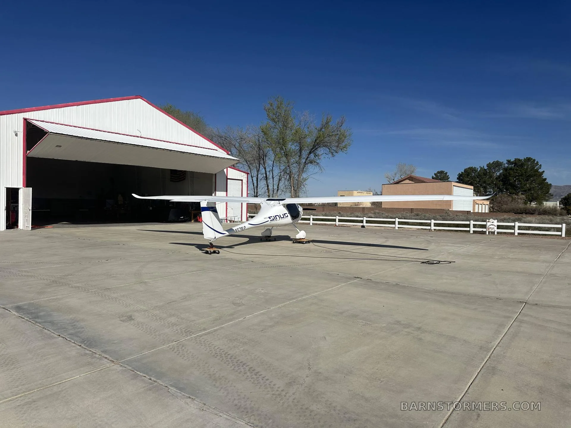 A white Pipistrel Sinus aircraft with blue accents is viewed from the rear-left, parked on an apron in front of a hangar.