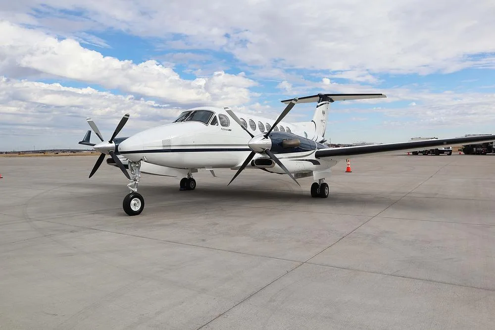A white and dark blue Beechcraft King Air 360 turboprop aircraft parked on an airport tarmac under a cloudy sky.