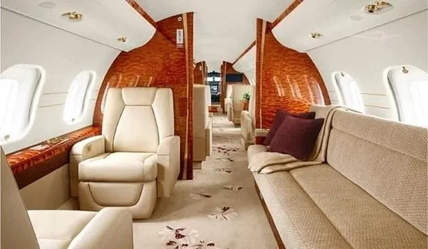 A view of a private jet's luxurious cabin, showing a cream sofa, individual seats, and wood paneling.