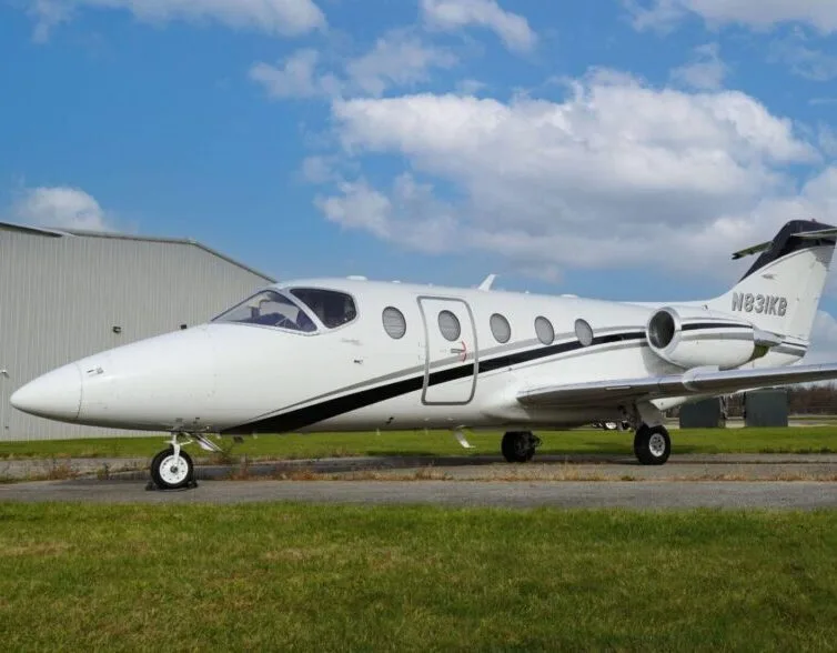 A white business jet with black and silver stripes, registration N831KB, parked on an airfield next to a hangar under a partly cloudy sky.