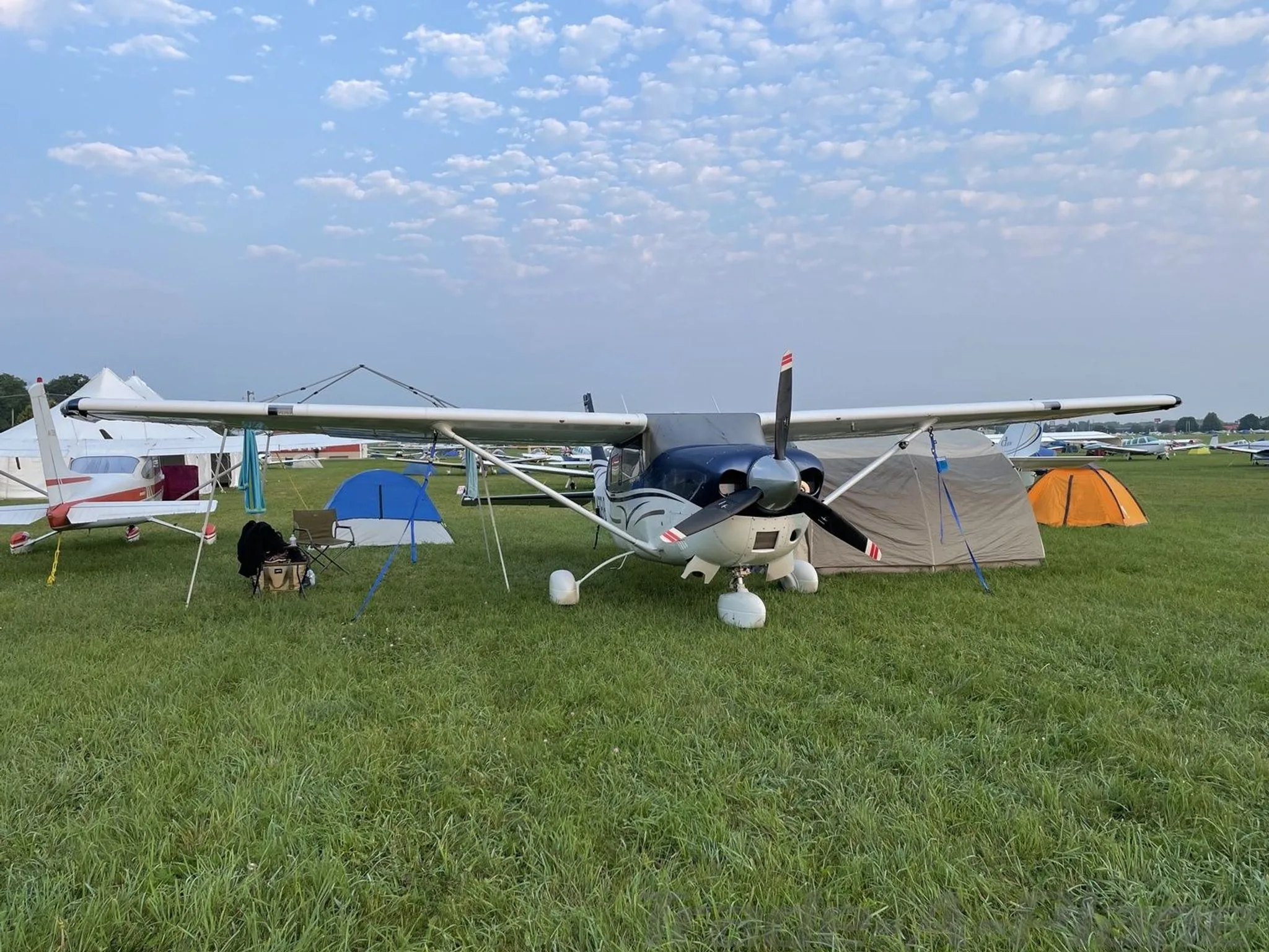 A blue and white Cessna 182 aircraft parked on a grassy field with camping tents under a cloudy sky.