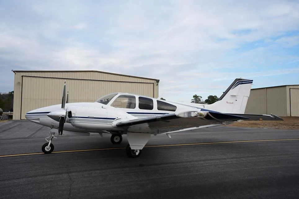 A white twin-engine propeller aircraft with blue and grey stripes parked on an asphalt ramp next to hangars.