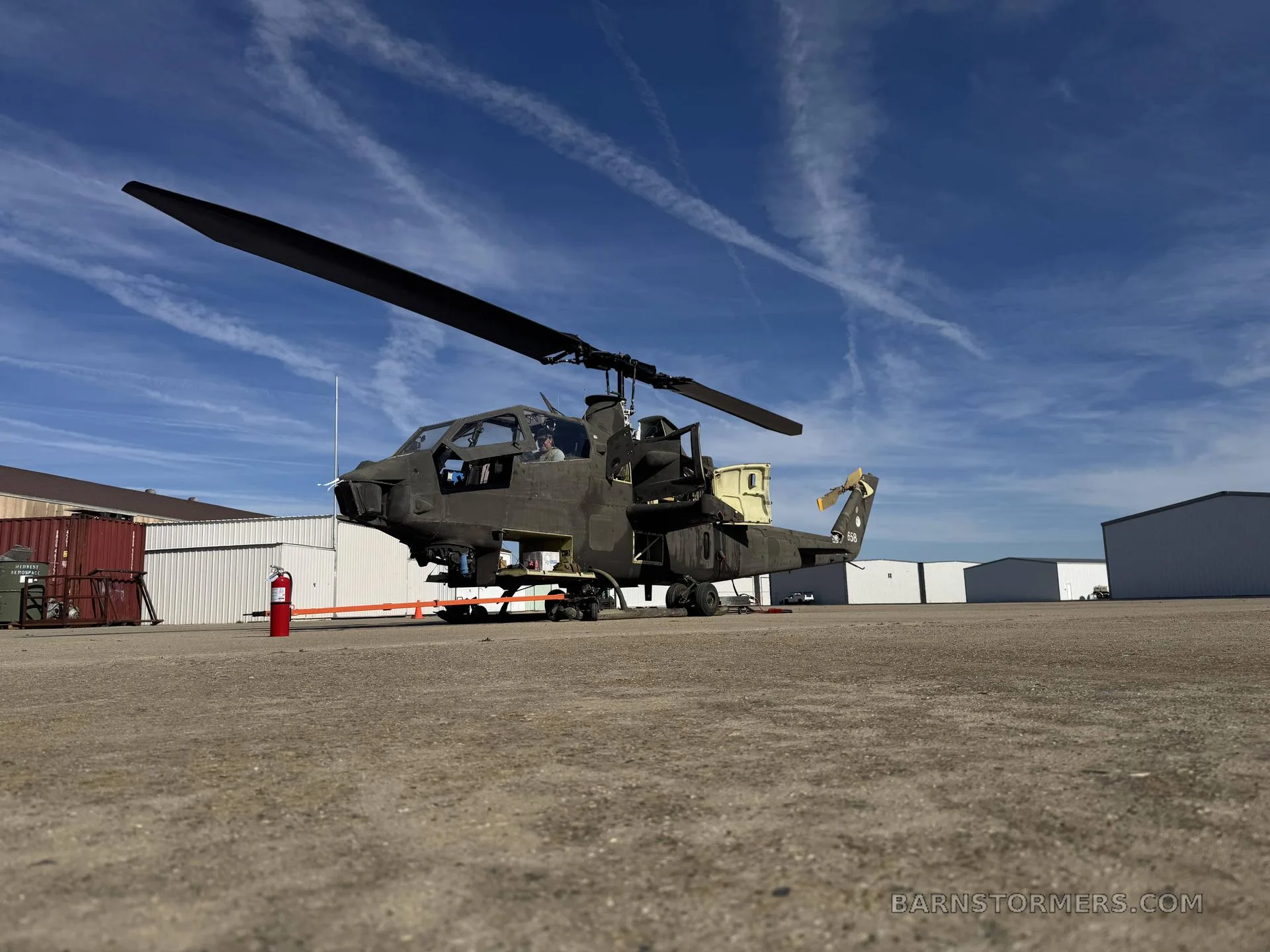 A partially disassembled dark green military helicopter, likely an Apache, sits on an airfield under a blue sky with contrails.