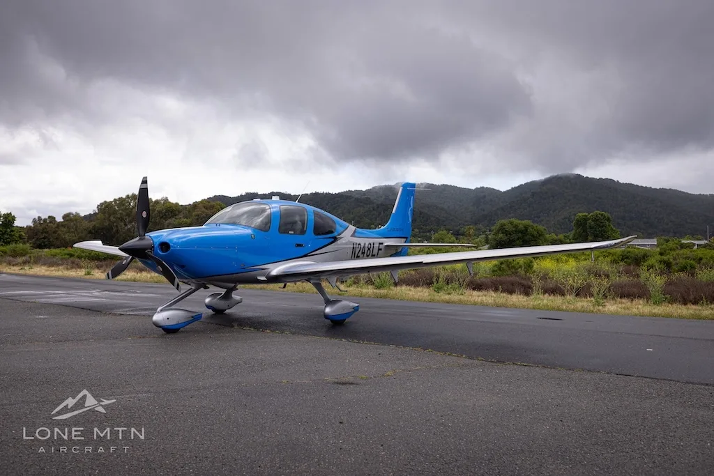 A blue and silver single-engine propeller aircraft (Cirrus SR22) with registration N248LF parked on a wet tarmac under a cloudy sky.